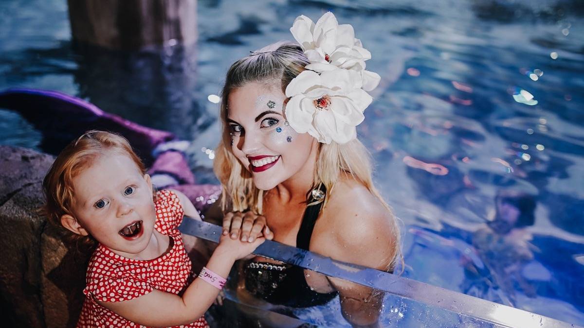 Small girl standing next to a mermaid in the water with a big white flower in her hair